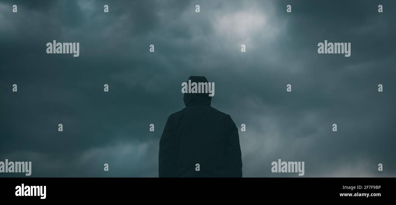 Rear view of male person wearing hooded jacket against dark moody dramatic clouds at sky, man looking into uncertain ominous future Stock Photo