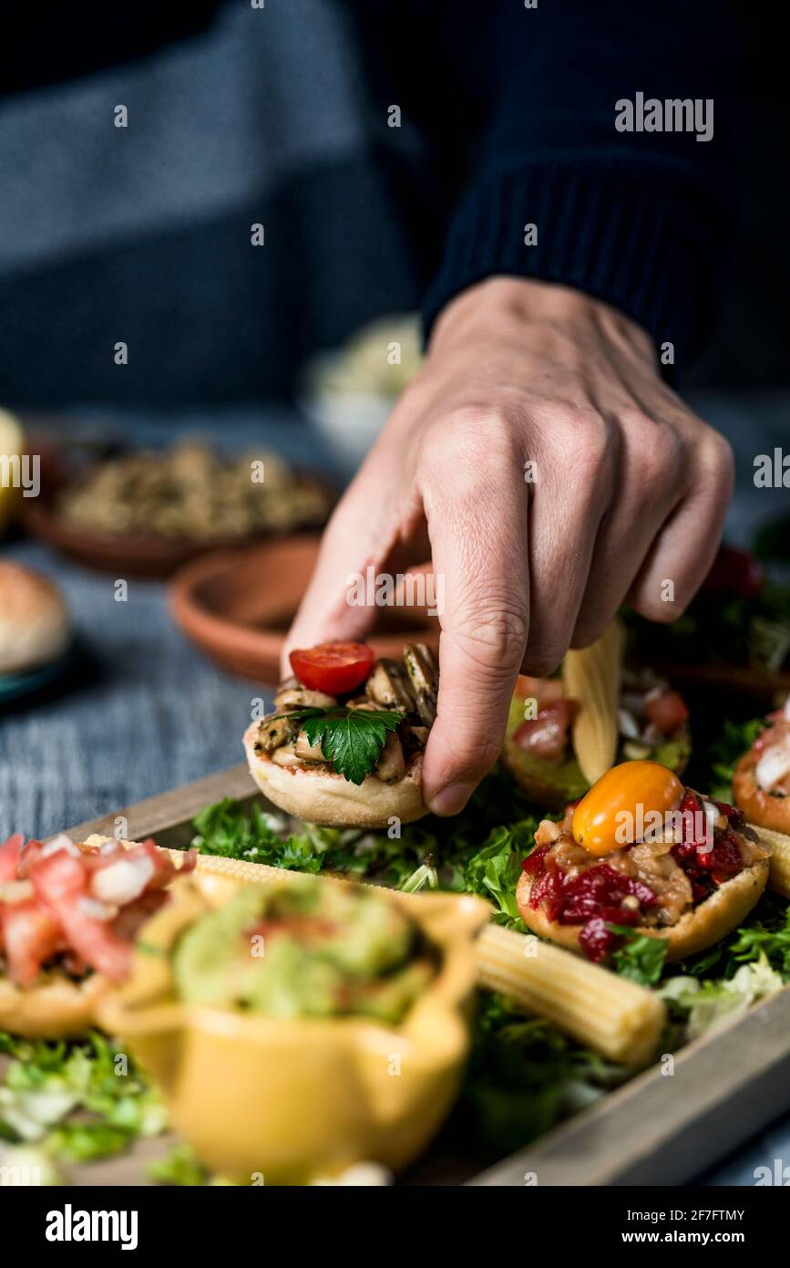 a young caucaisan man takes an appetizer from a wooden tray with some different vegan appetizers, with different toppings, placed on a gray rustic tab Stock Photo