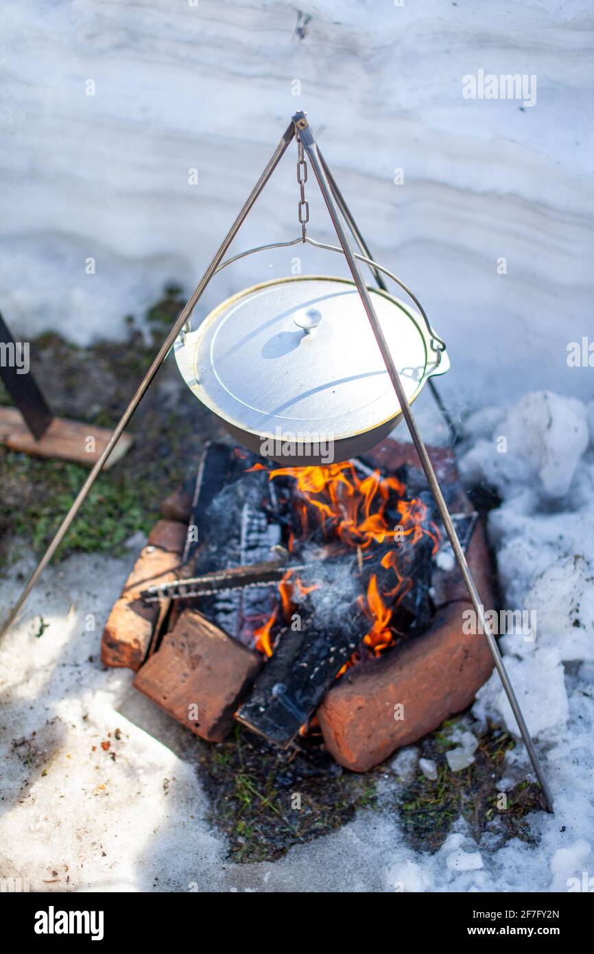 Over the fire hangs a pot in which to cook food. On a hook on a tripod,  steam comes out of the pan. Winter Camping outdoor cooking Stock Photo -  Alamy