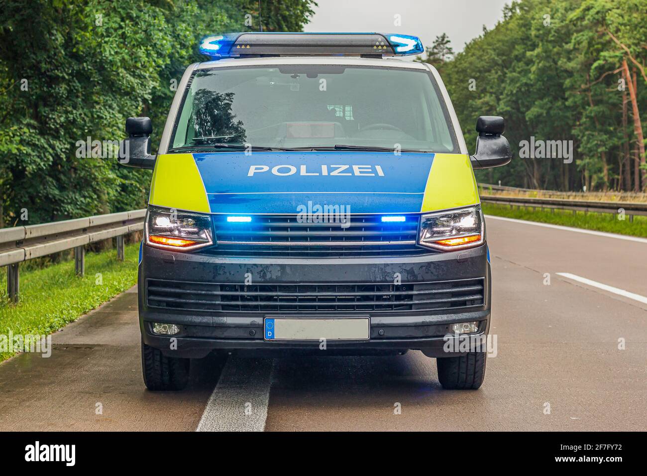 Front view of a police car on the highway. Switched on blue light in the headlights of the police car. Rainy weather in spring with green trees. Stock Photo