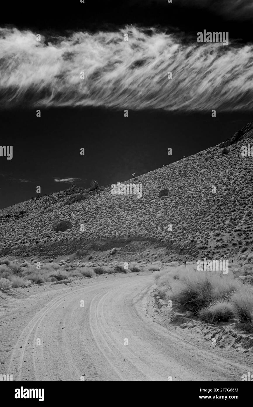 Lonely dirt road leading towards desert hills under black sky with white clouds. Monochrome image. Stock Photo
