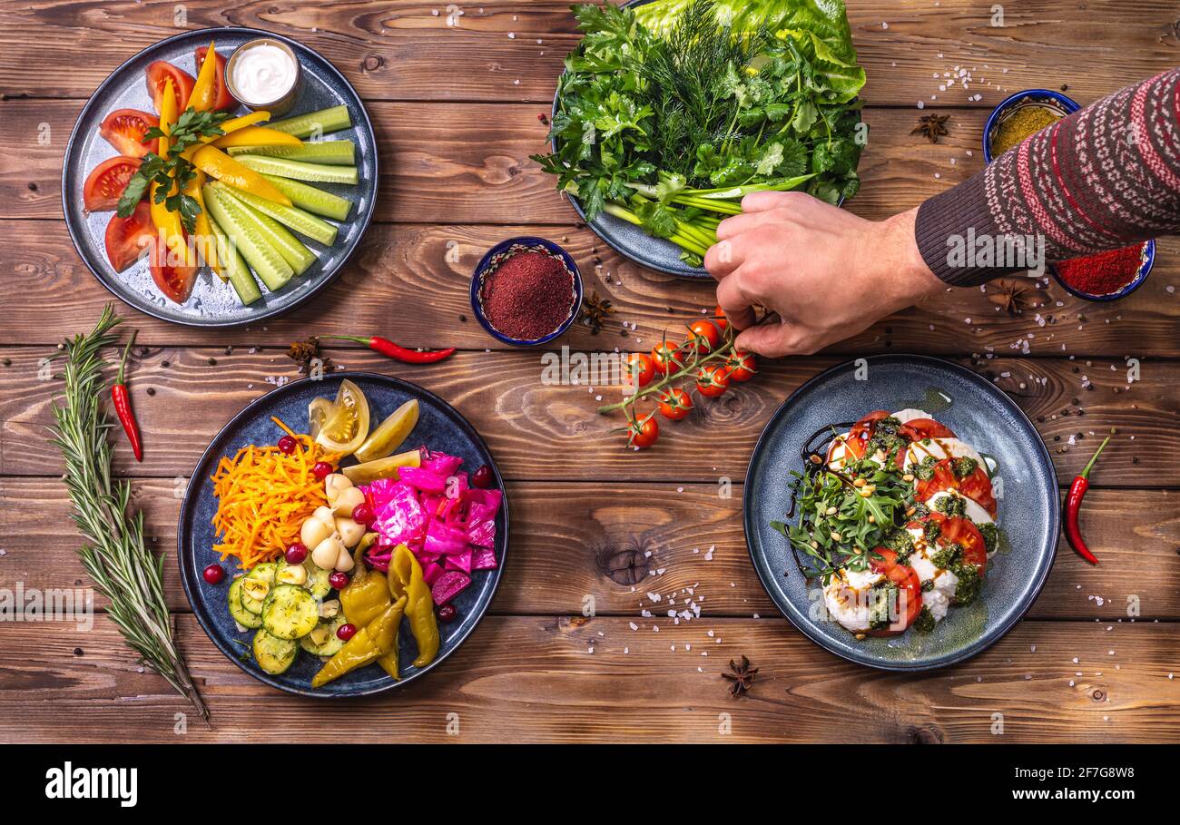Pickled vegetables: peppers, carrots, garlic, cabbage lie on a platter on a wooden background decorated with chili peppers. The cook's hand puts cherr Stock Photo