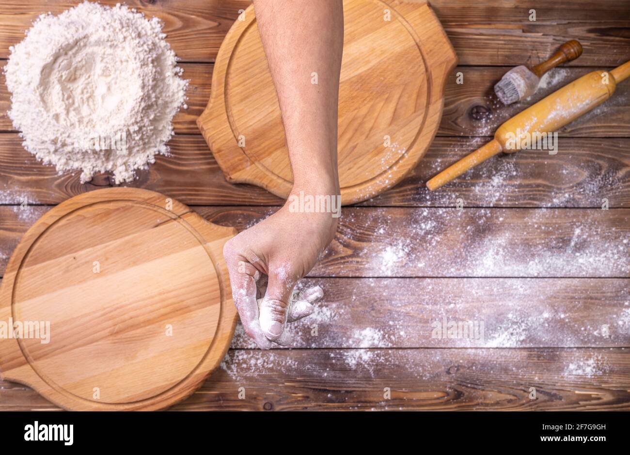 The chef's hand sprinkles wheat flour on wooden round pizza boards, chicken eggs, and a rolling pin on a brown board background. Selective focus. Prep Stock Photo