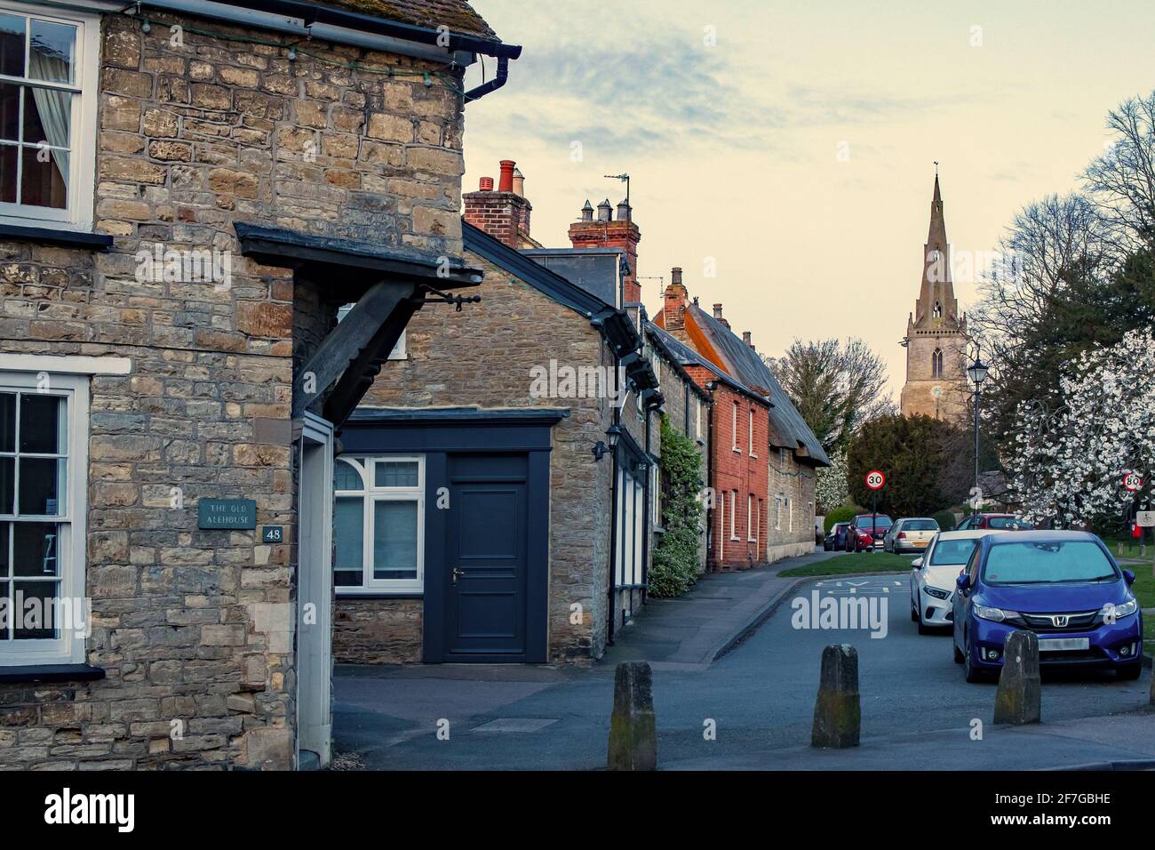 Church Lane, Sharnbrook, Bedfordshire, England, UK - springtime early morning view along village street with parked cars and church tower in distance Stock Photo