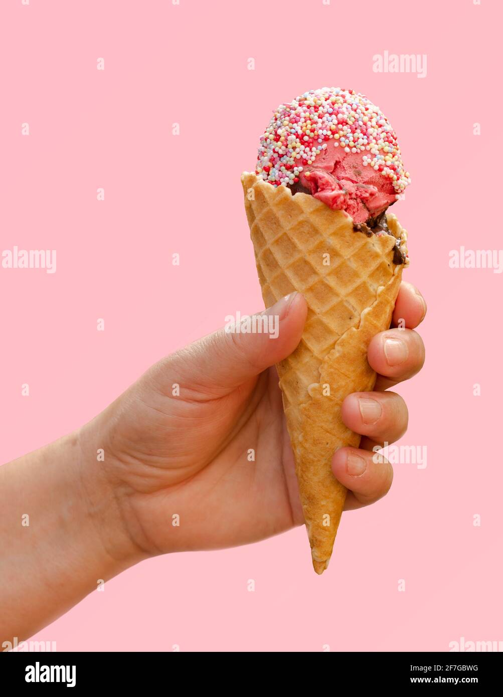 Hand holding strawberry ice cream in waffle cone with candy sprinkles isolated on light pink background, taste of summer concept, frozen dessert Stock Photo