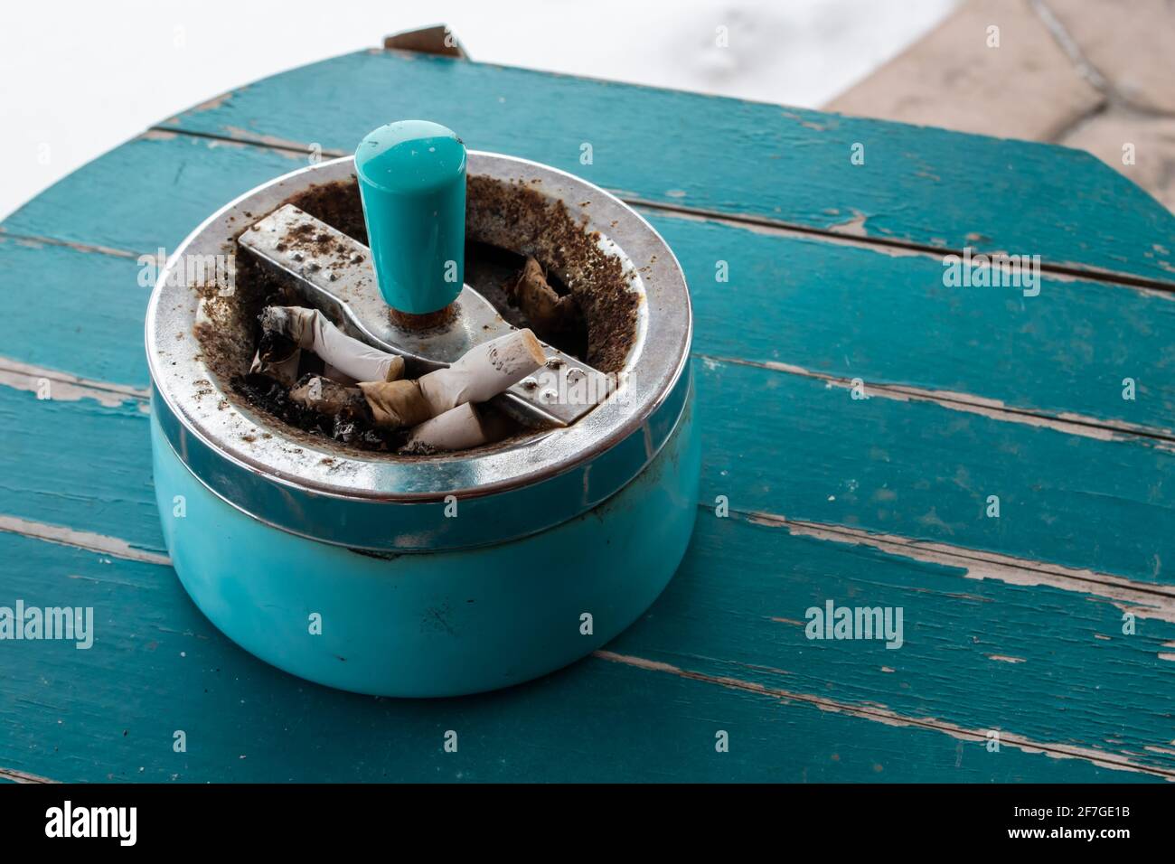 A retro, antique cigarette ashtray filled with stubs on a turquoise sanded wooden table top. Angle side view. Stock Photo