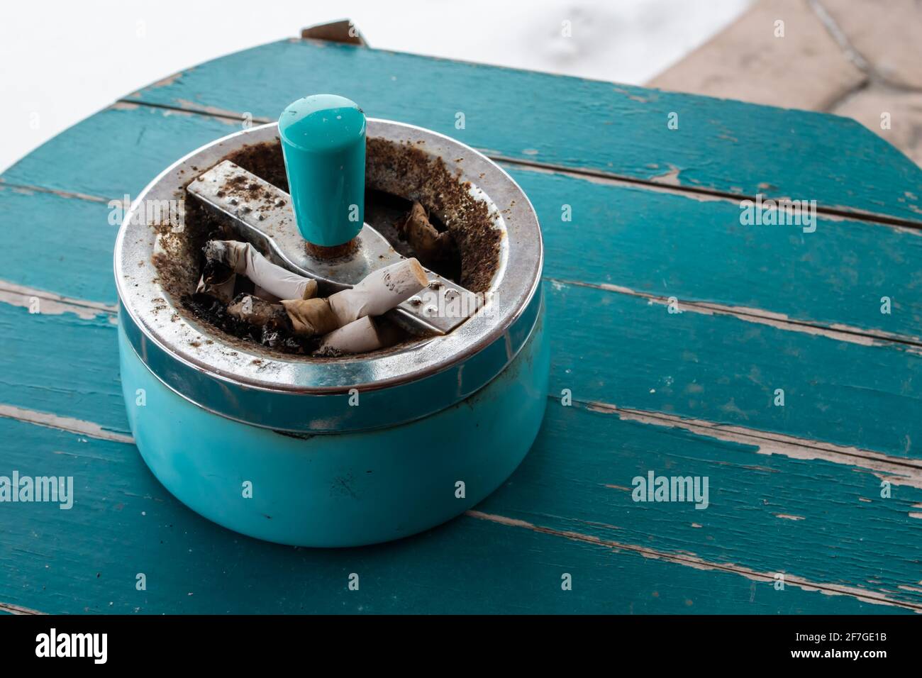 A retro, antique cigarette ashtray filled with stubs on a turquoise sanded  wooden table top. Angle side view Stock Photo
