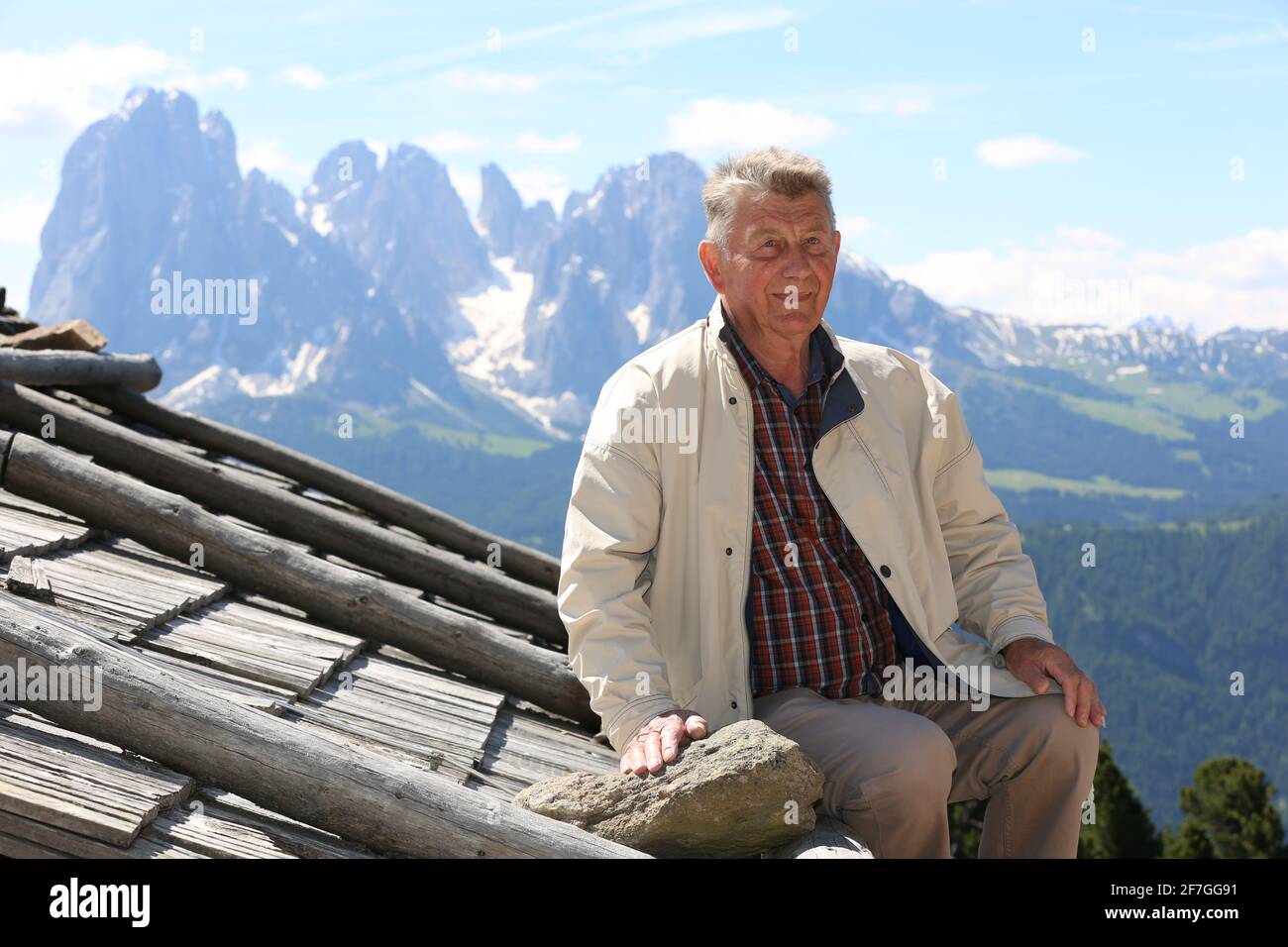 sportlicher Senior auf dem Holzdach einer südtiroler Almhütte vor dem Panorama von Langkofel und Plattkofel in der Abendsonne der Dolomiten in Italien Stock Photo