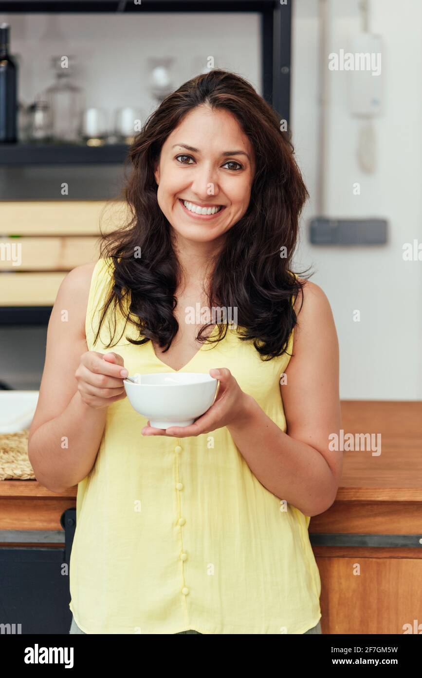 Cheerful adult female eating healthy food and looking at camera while leaning on table during breakfast in kitchen at home Stock Photo