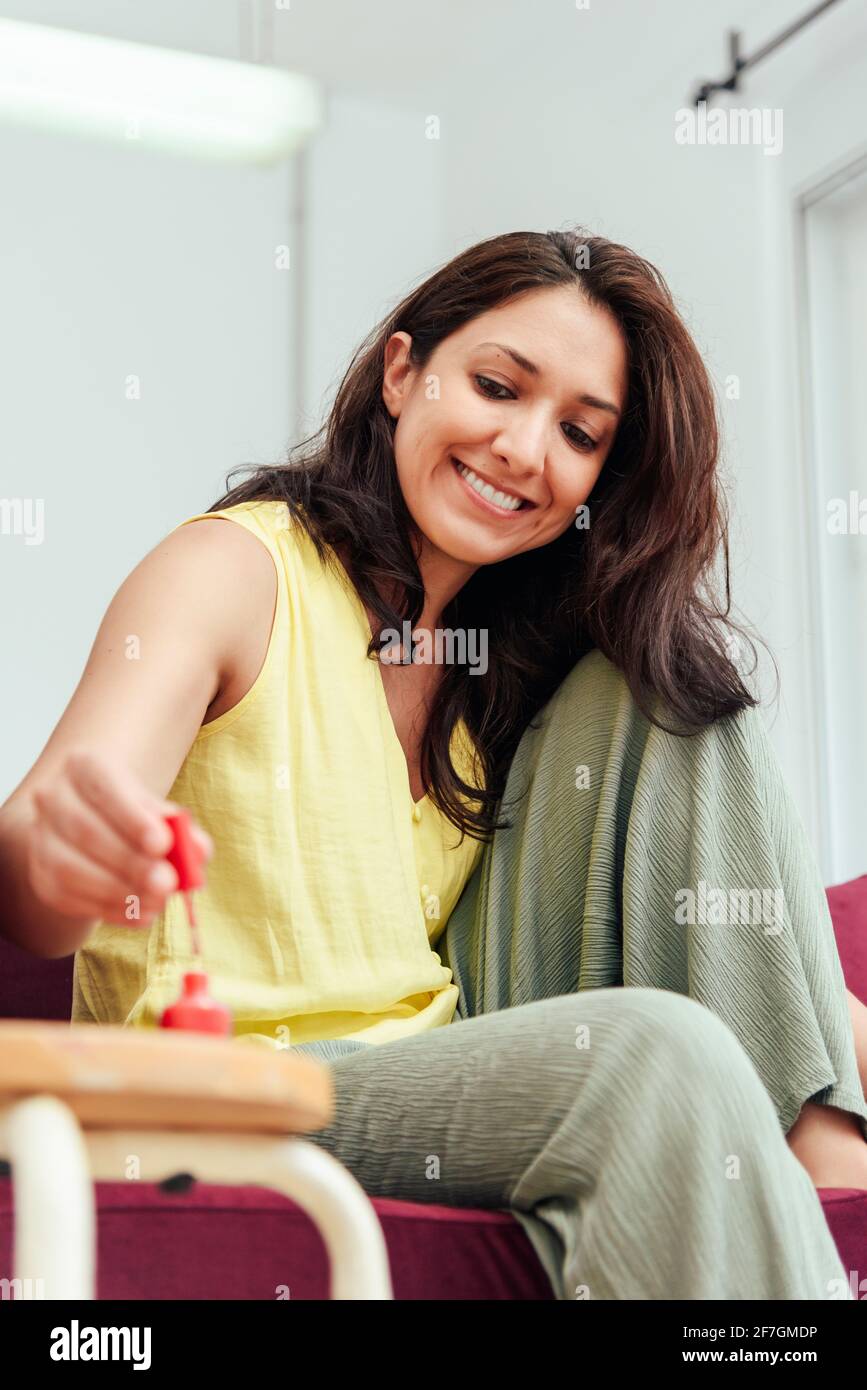 Happy adult woman smiling and applying nail polish on toenails while sitting on sofa on weekend day at home Stock Photo