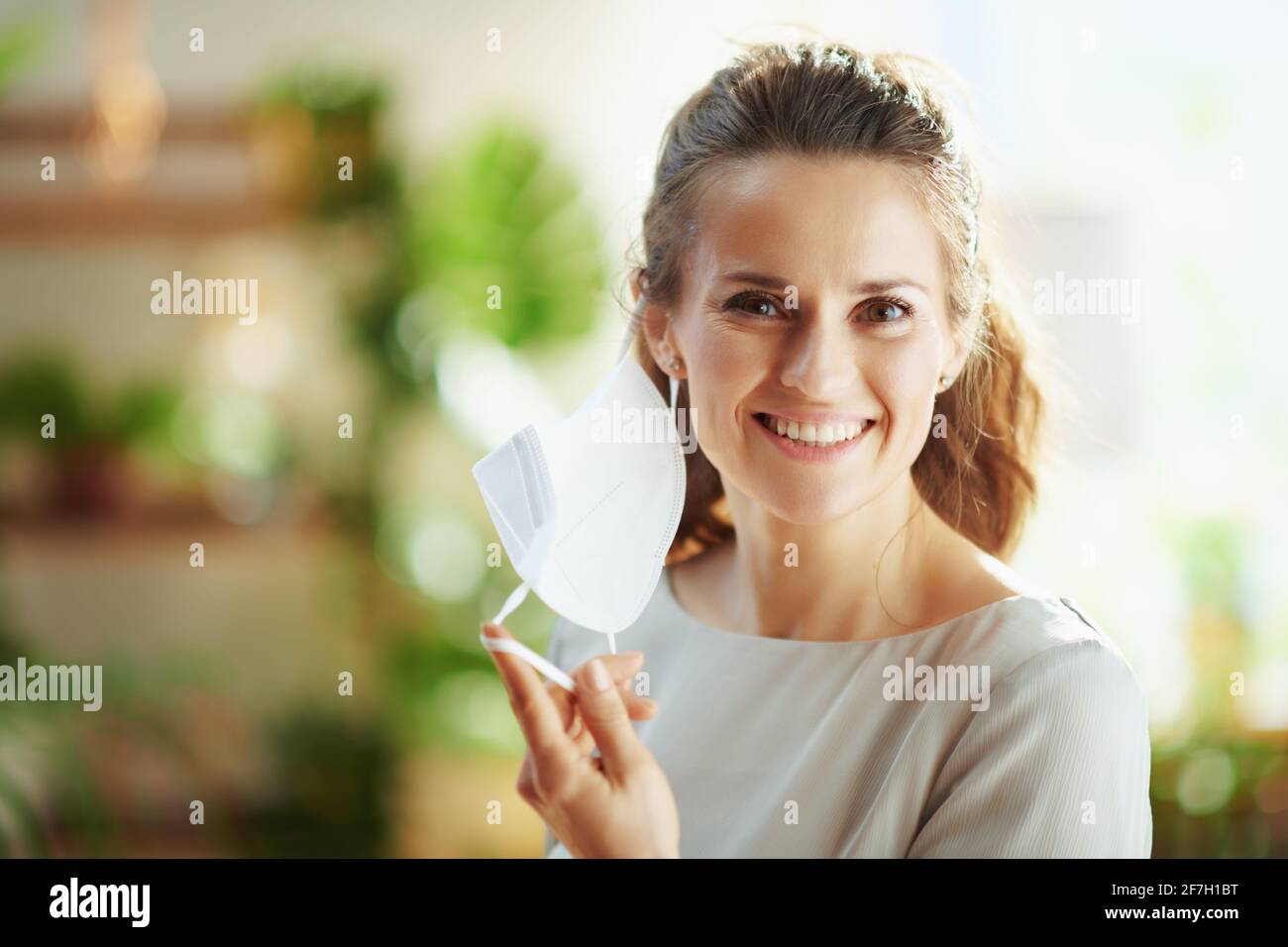 covid-19 pandemic. Portrait of smiling elegant woman in grey blouse taking off ffp2 mask. Stock Photo