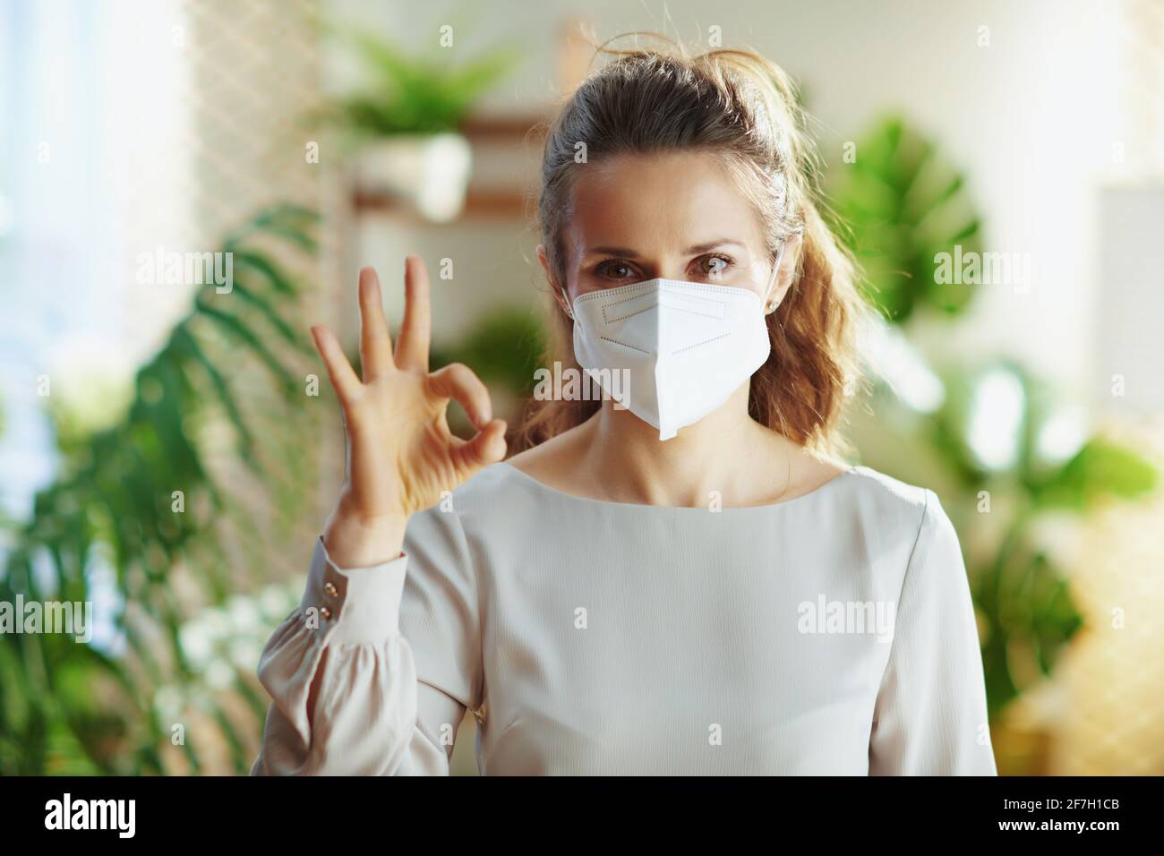 coronavirus pandemic. smiling trendy middle aged woman in grey blouse with ffp2 mask in the modern house showing ok gesture. Stock Photo