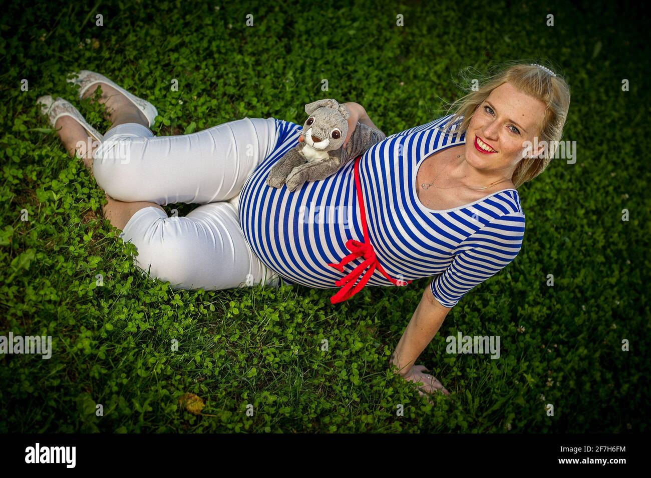 A cute pregnant girl with a striped top sitting in a green grass holding a fluffy bunny toy. Stock Photo