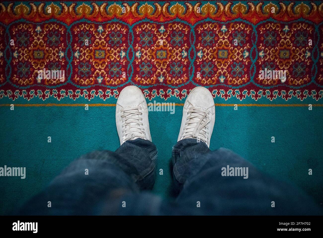 Foot of a man standing on a hotel floor mat or rug, wearing jeans and white sneakers. View from above. Stock Photo