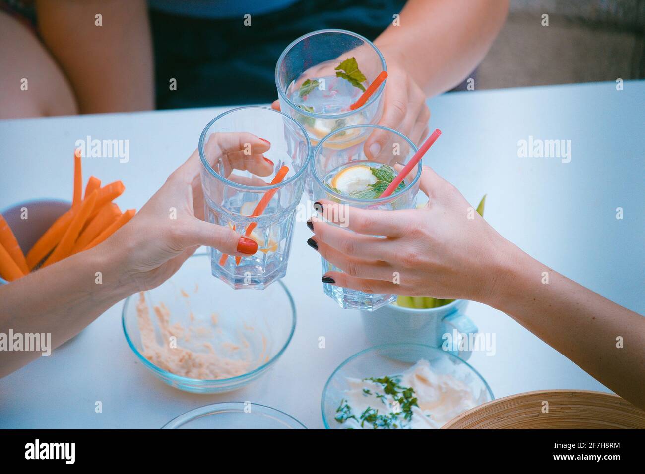 Hands of three women viewed from above seen cheering or toasting with glasses of gin tonic drink. Other healthy foods are seen around, like dips and c Stock Photo