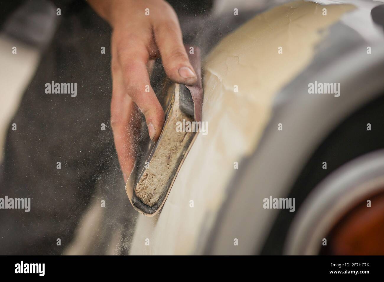 A hand of a man is seen sanding an old vintage car using a block of wood and brown sand grit paper. Manual dry sanding of a car in restoration, a proc Stock Photo