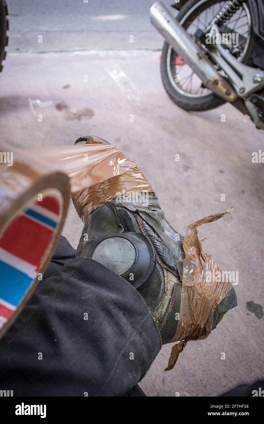 A person taping up his broken or used leather motorcycle boots with brown tape or sellotape in order to lengthen their lifespan. Motorcycle in the bac Stock Photo