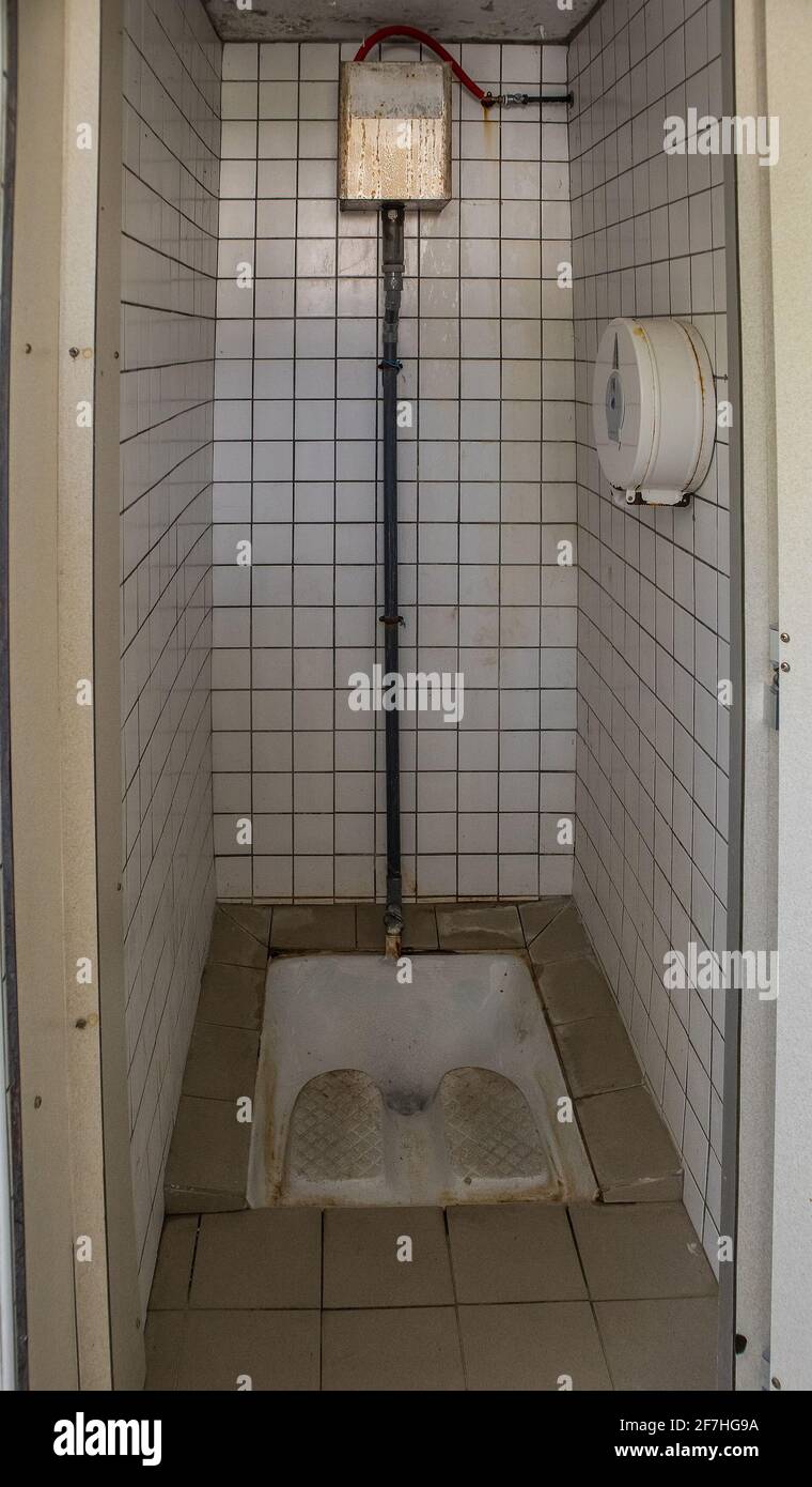 A public squat toilet with white tiles and other ceramic parts. Visible toilet paper and water container on the wall. Dirty, but not totally dirty and Stock Photo
