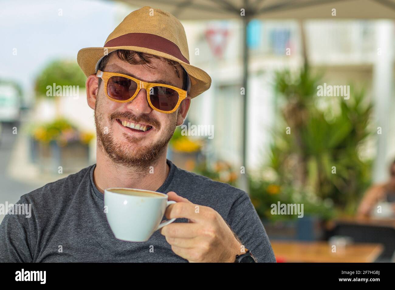 Hipster looking man with thatched hat and wooden sunglasses holding a white cup of coffee while drinking in smiling and sitting in a cafe environment. Stock Photo