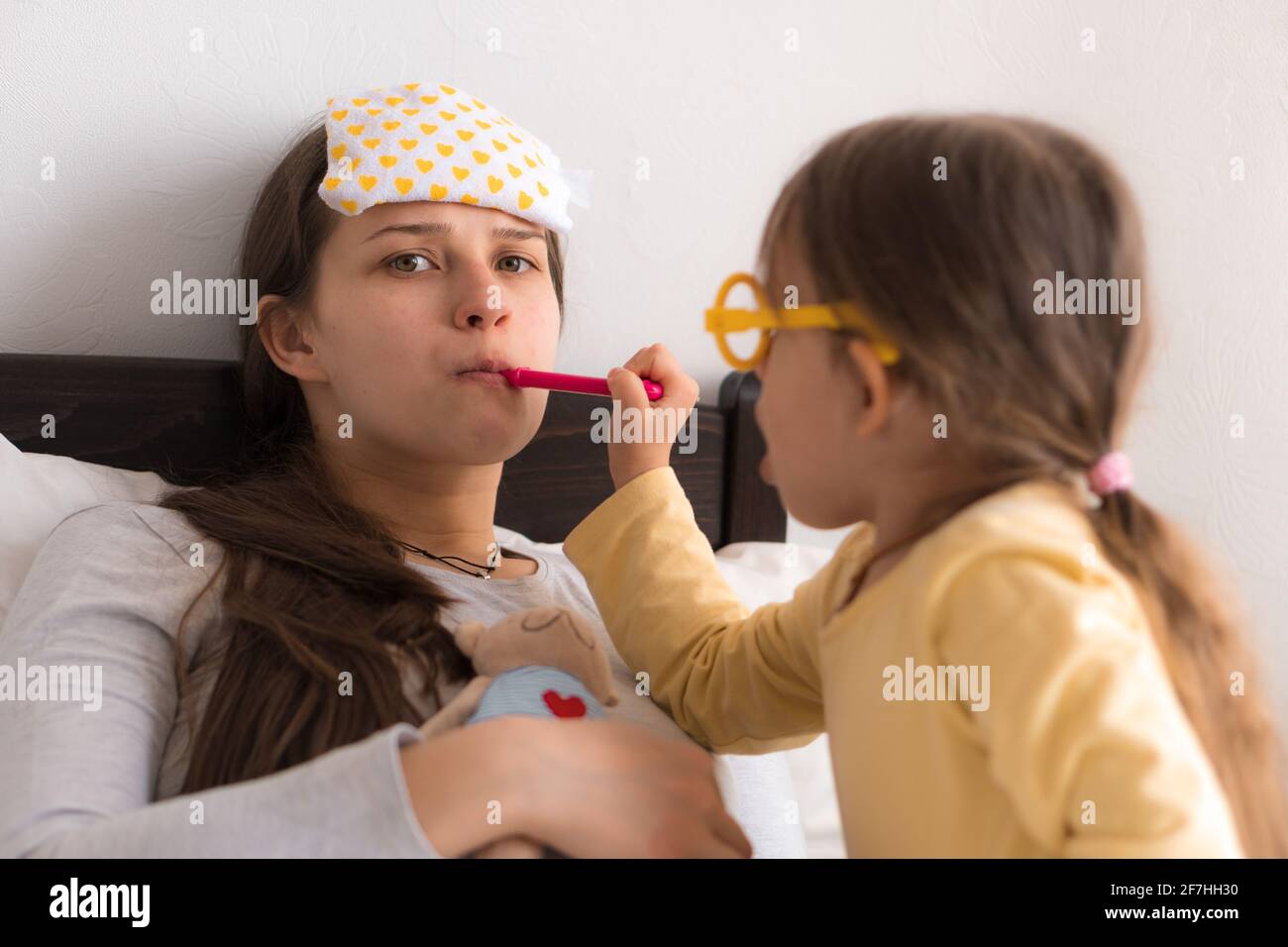 Cute little girl wearing uniform playing doctor or nurse with young mum or nanny in bedroom, checking mother throat, measures temperature, family Stock Photo