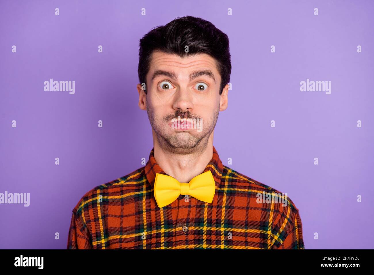Photo of crazy foolish guy puffed cheeks look camera wear bow tie checkered shirt isolated violet background Stock Photo