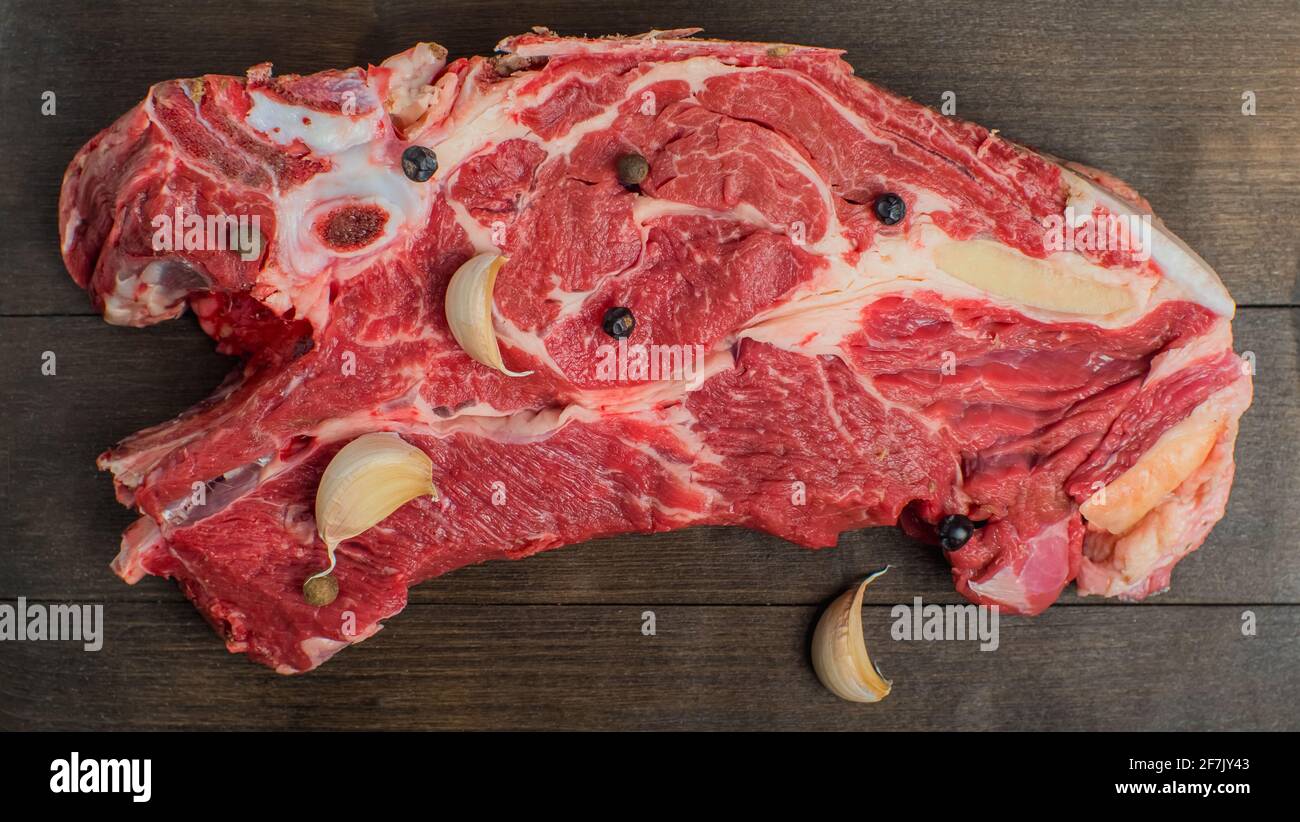 A piece of fresh red beef with cloves of garlic and peas of pepper lies on a dark cutting board. Close-up Stock Photo