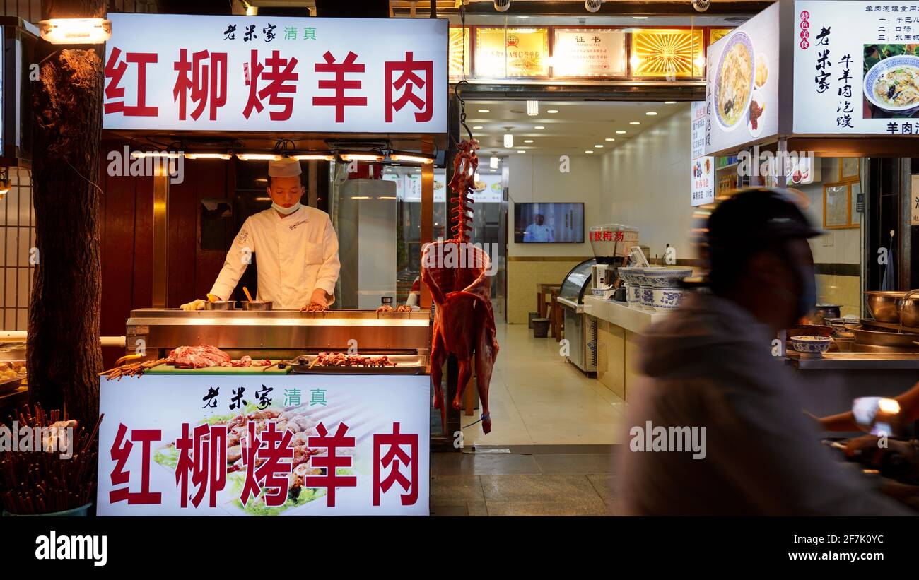 A young man is cooking lamb kebab in a booth as traditional Chinese food, with another person passing by. Stock Photo