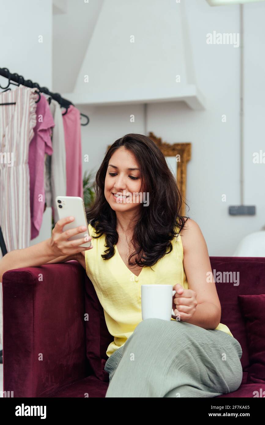 Young woman sitting on the sofa in her apartment drinking coffee while checking her social networks. Stock Photo