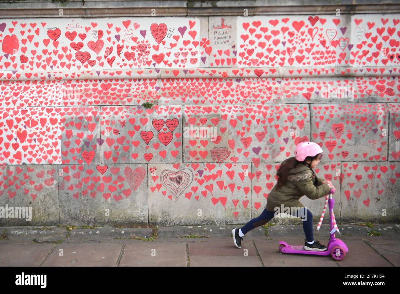 A young girl rides a scooter past the National Covid Memorial Wall on the Embankment in London. More than one thousand people, including the bereaved, NHS staff, volunteers and members of the public have been painting 150,000 hearts onto the wall, which now stretches for around half a kilometre, to represent loved ones lost to the virus in the UK. Picture date: Thursday April 8, 2021. Stock Photo