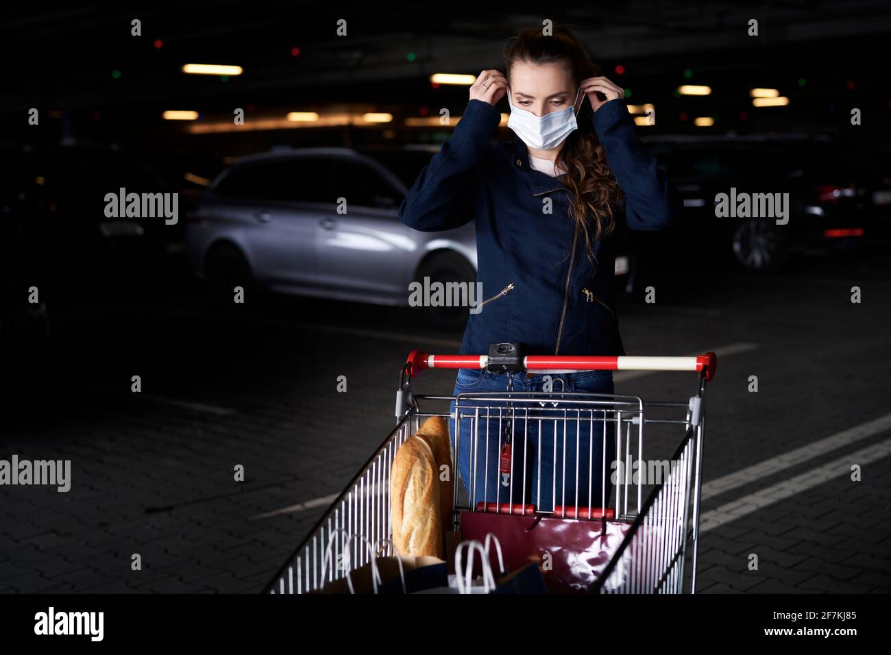 Young woman with shopping cart assumes protective mask on parking Stock Photo