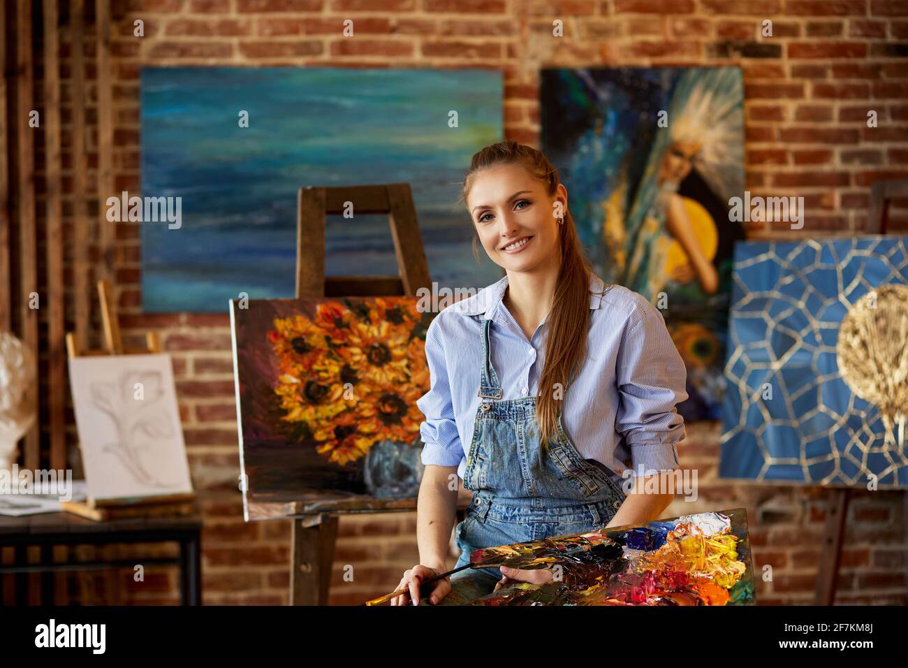 Portrait of young smiling female artist with her artworks in art studio ...
