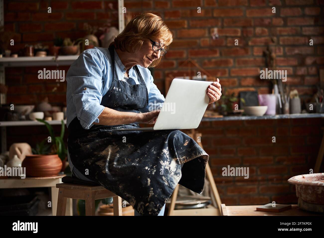 Charming old woman using modern laptop in pottery workshop Stock Photo