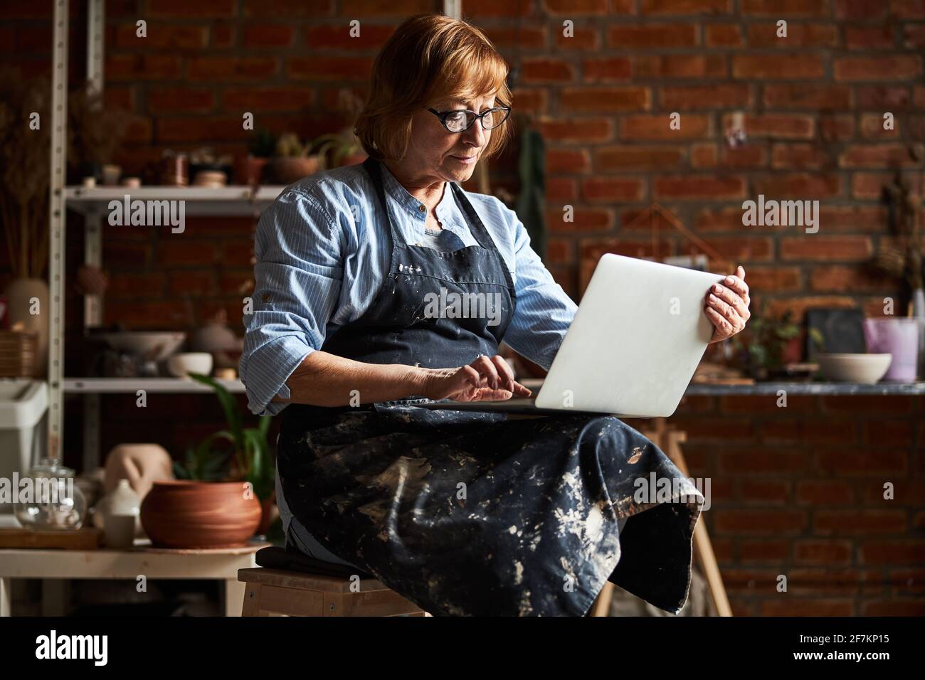 Nice old woman using modern laptop in pottery workshop Stock Photo