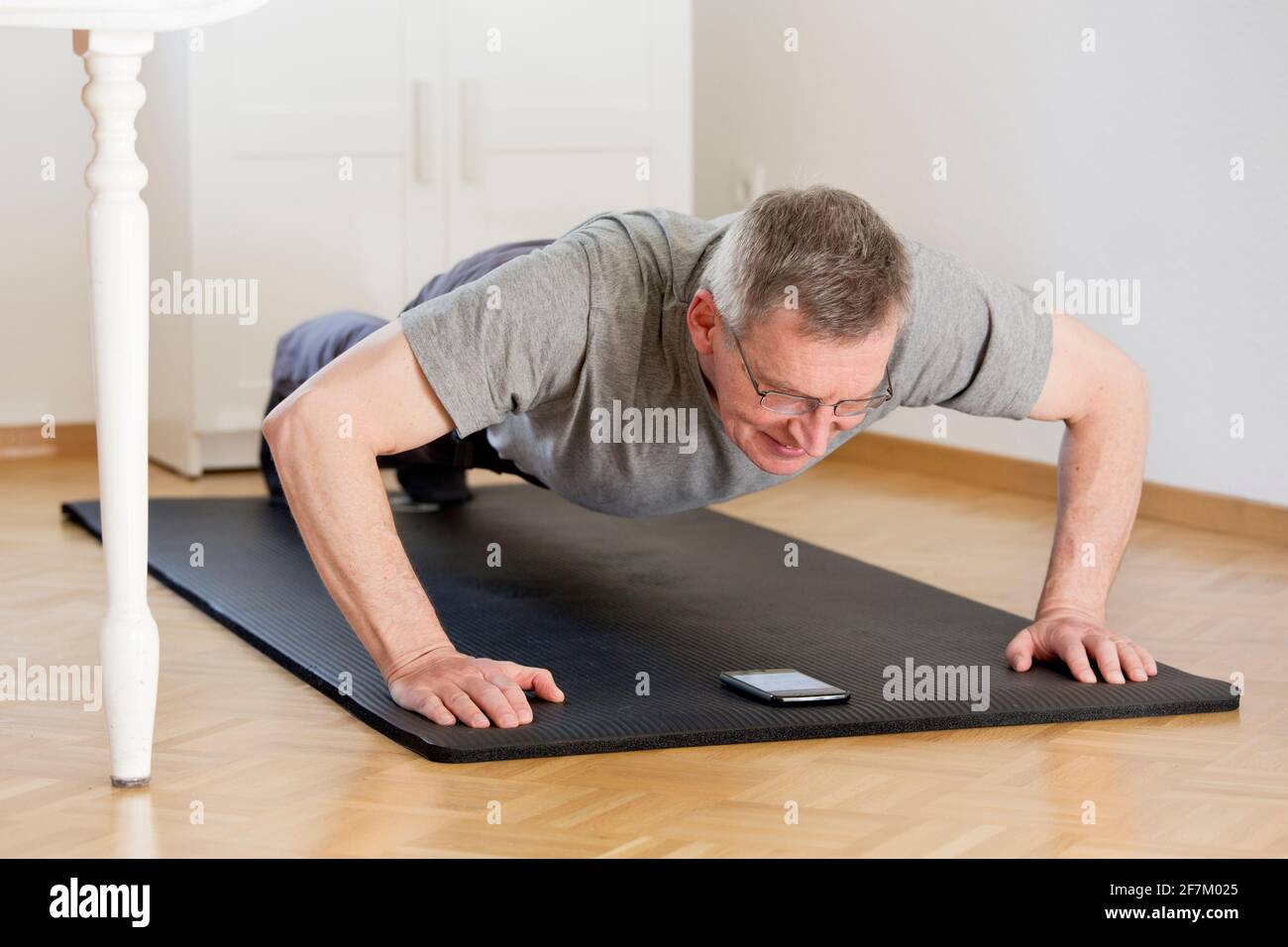 Mature man doing push ups at home with smart phone on the mat - selective focus on the face Stock Photo