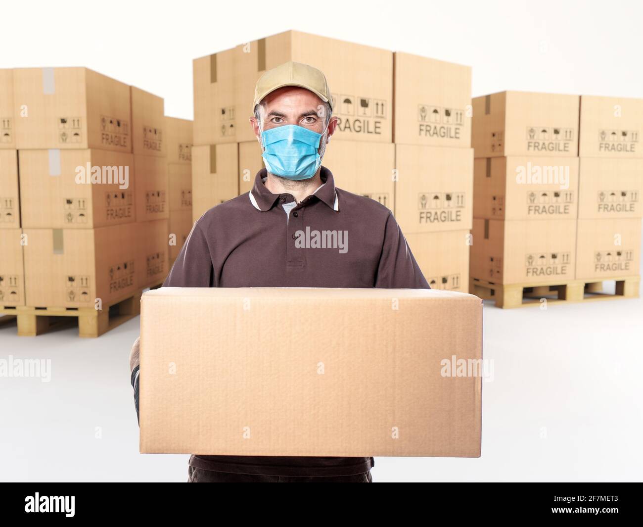 courier with mask and parcel in hand, pallet of cartons in the background. Stock Photo