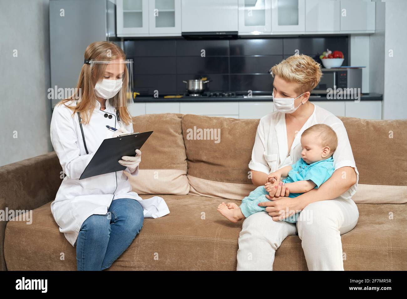 Pleasant woman in medical mask sitting on couch with her little son on knees while female doctor writing on clipboard near. Health care for children at home. Stock Photo