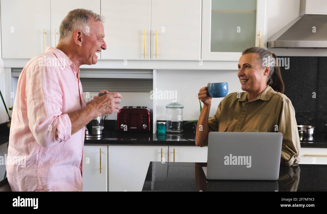 Happy caucasian senior couple in kitchen drinking coffee and laughing, woman using laptop Stock Photo
