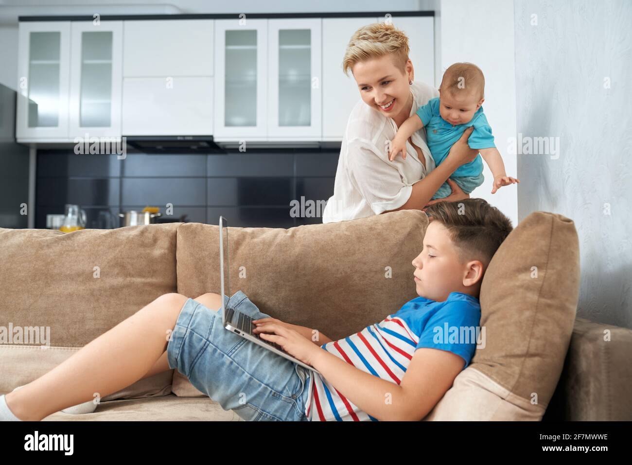 Pretty boy lying on couch with laptop on knees, mother with little baby on hands standing near and looking at computer screen. Concept of family and lifestyle. Stock Photo