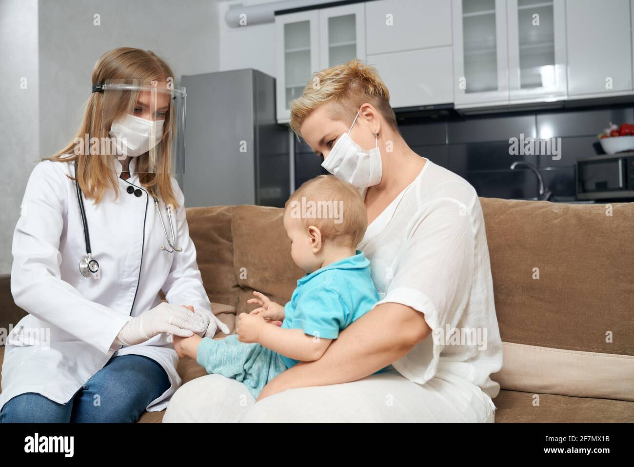 Professional doctor in medical uniform and mask checking health condition of little baby. Young mother holding boy on knees during medical examination. Stock Photo
