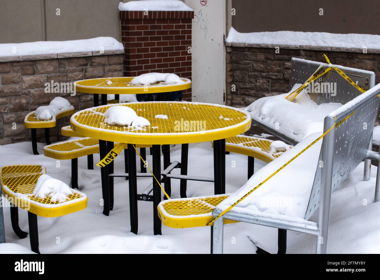 London, Ontario, Canada - February 15 2021: A yellow metal grill table with benches outside a Marble slab creamery in London sits covered in snow. Stock Photo