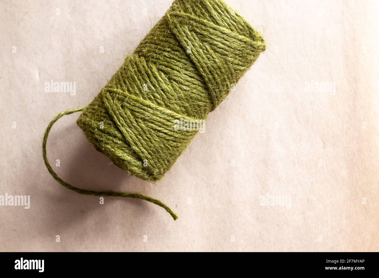 A cylinder of olive green yarn with a single strand hanging loose on a parchment paper background in natural light, Ontario, Canada, 2021. Stock Photo