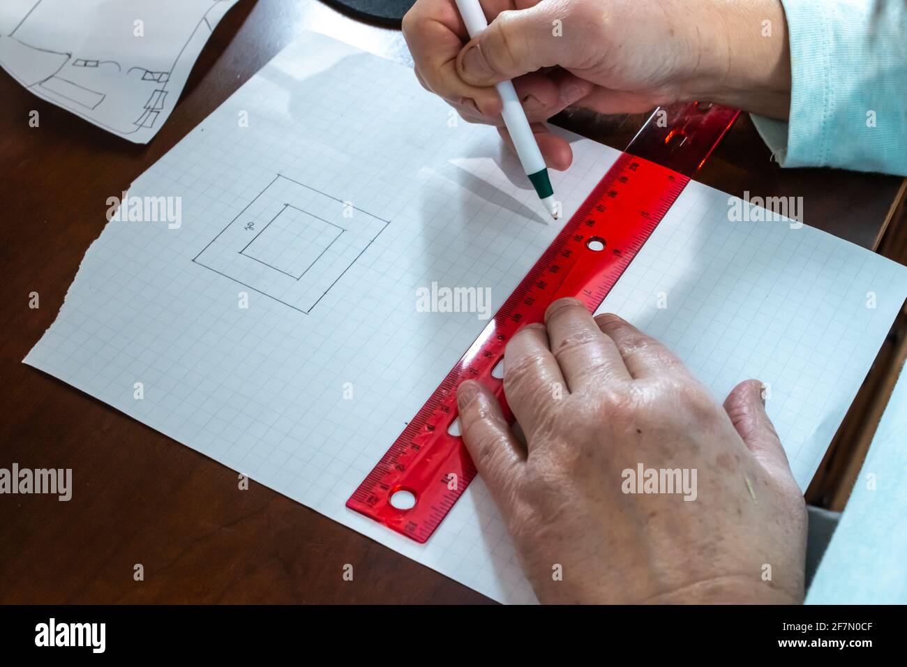 A woman uses a pen to count measurements and sketch out a floor plan for a house on square graph paper using a clear red ruler on hardwood desk. Stock Photo