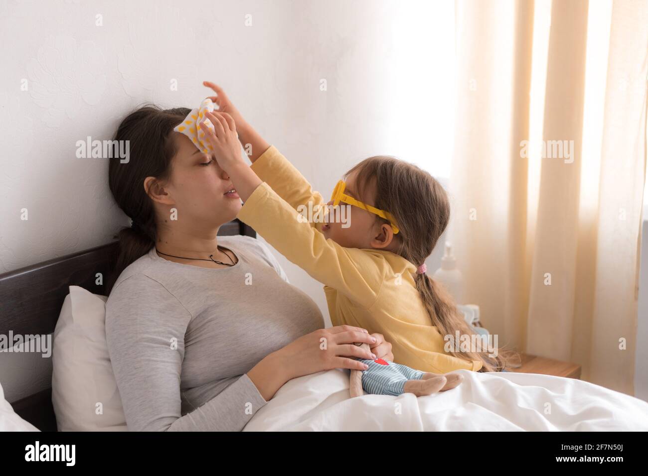 little girl wearing uniform playing doctor, nurse with young mum, nanny in bedroom, checking mother throat, measures temperature, take care, family Stock Photo