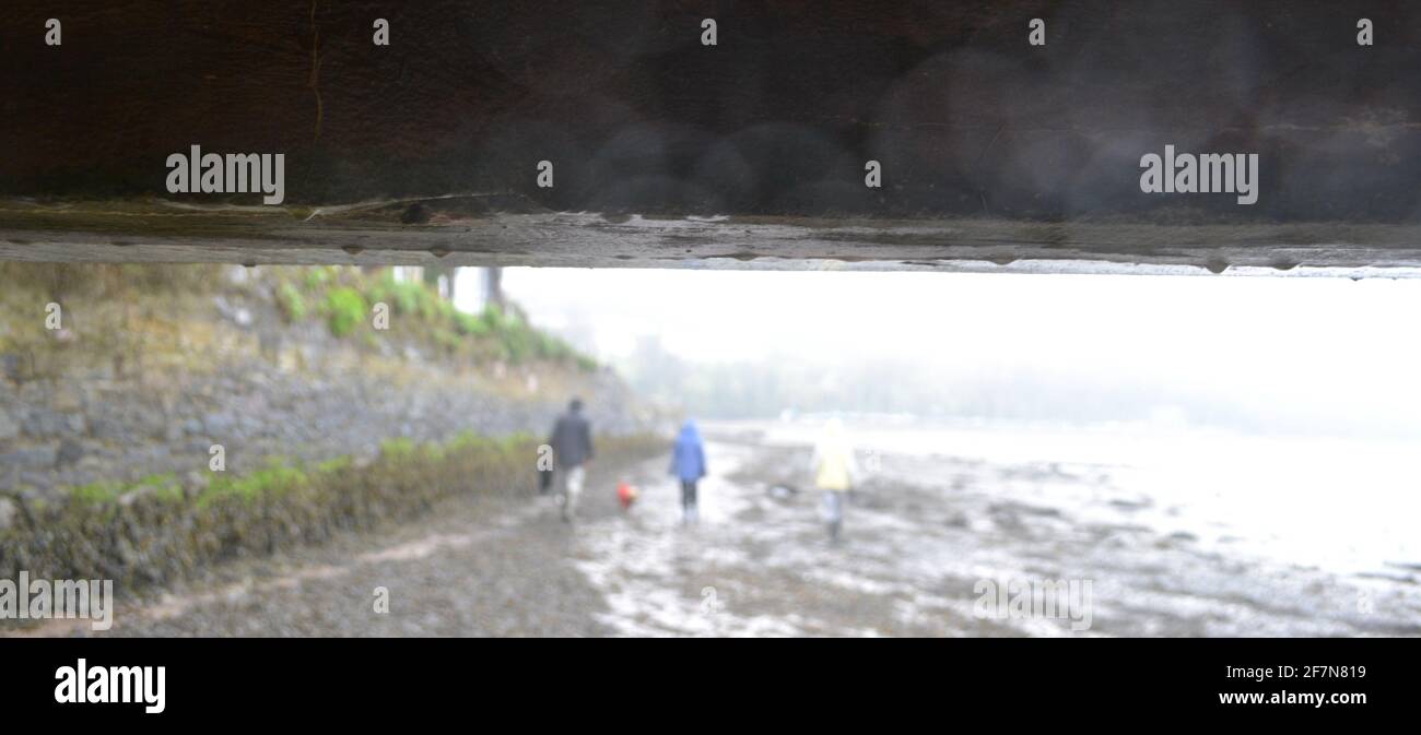 A damp wooden beam or bridge on a rainy day. Out of focus in the distance, three anonymous human figures, wearing waterproof raincoats, walk a dog Stock Photo
