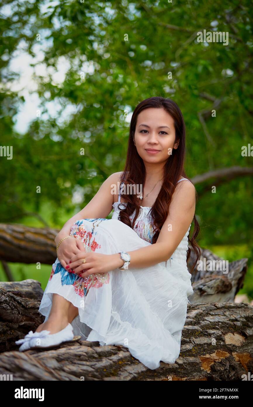Full body adult filipino female in dress looking at camera while relaxing on trunk of tree on summer day in forest Stock Photo