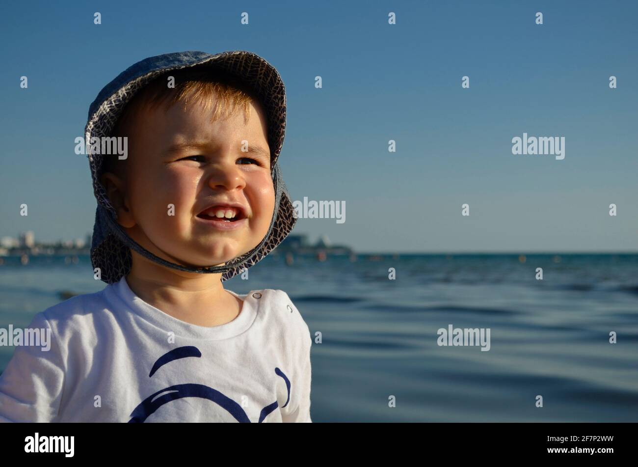 Emotional portrait of a child brightly lit. Looks concentrated, squints from the sun. Wearing a hat against the blue sea and sky, on the shore on a hot summer day. Stock Photo