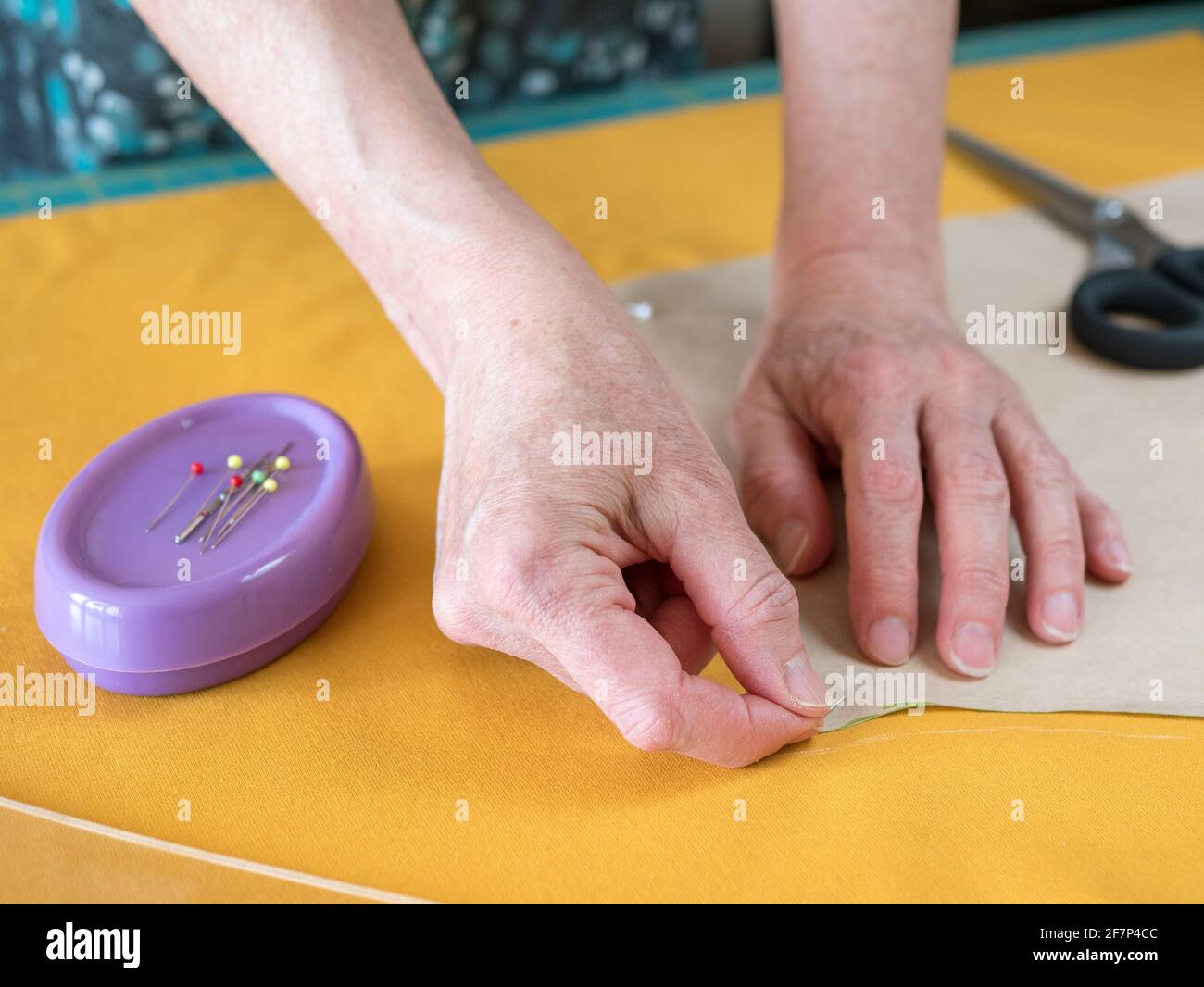 Hands of elderly woman are fixing a sewing pattern on the yellow fabric with a pin. Tailor is making a dress Stock Photo