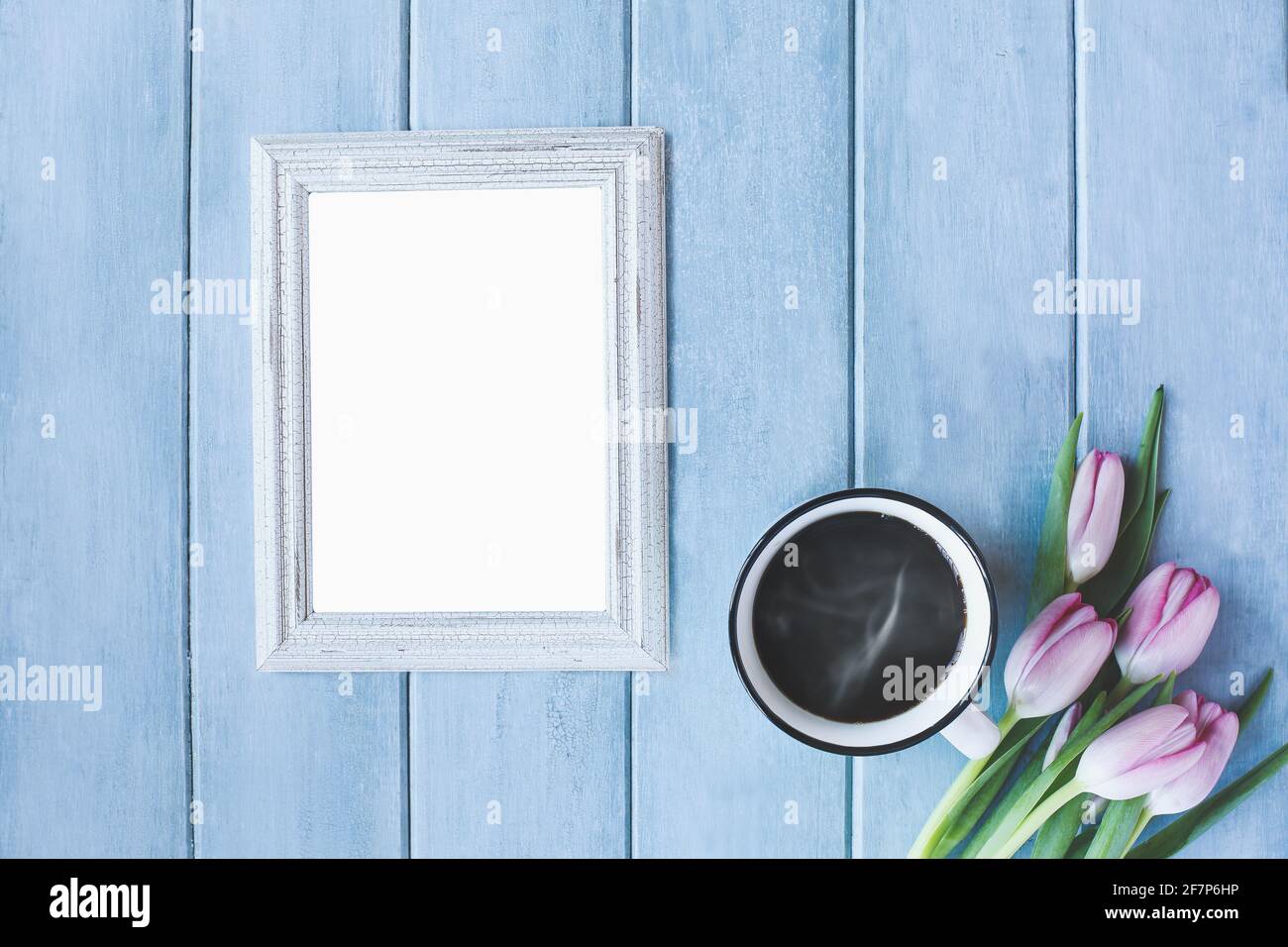 Vertical empty picture frame with steaming hot cup of black coffee and pink spring tulip flowers over a rustic blue wooden table shot from top view. Stock Photo
