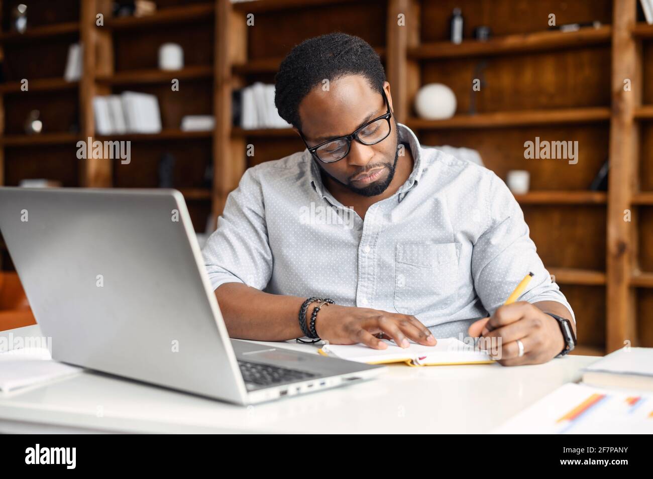 Focused African-American guy is using a laptop for watching webinars, taking notes, studying online. A male freelancer writes down startup ideas, sittin at the desk in office Stock Photo