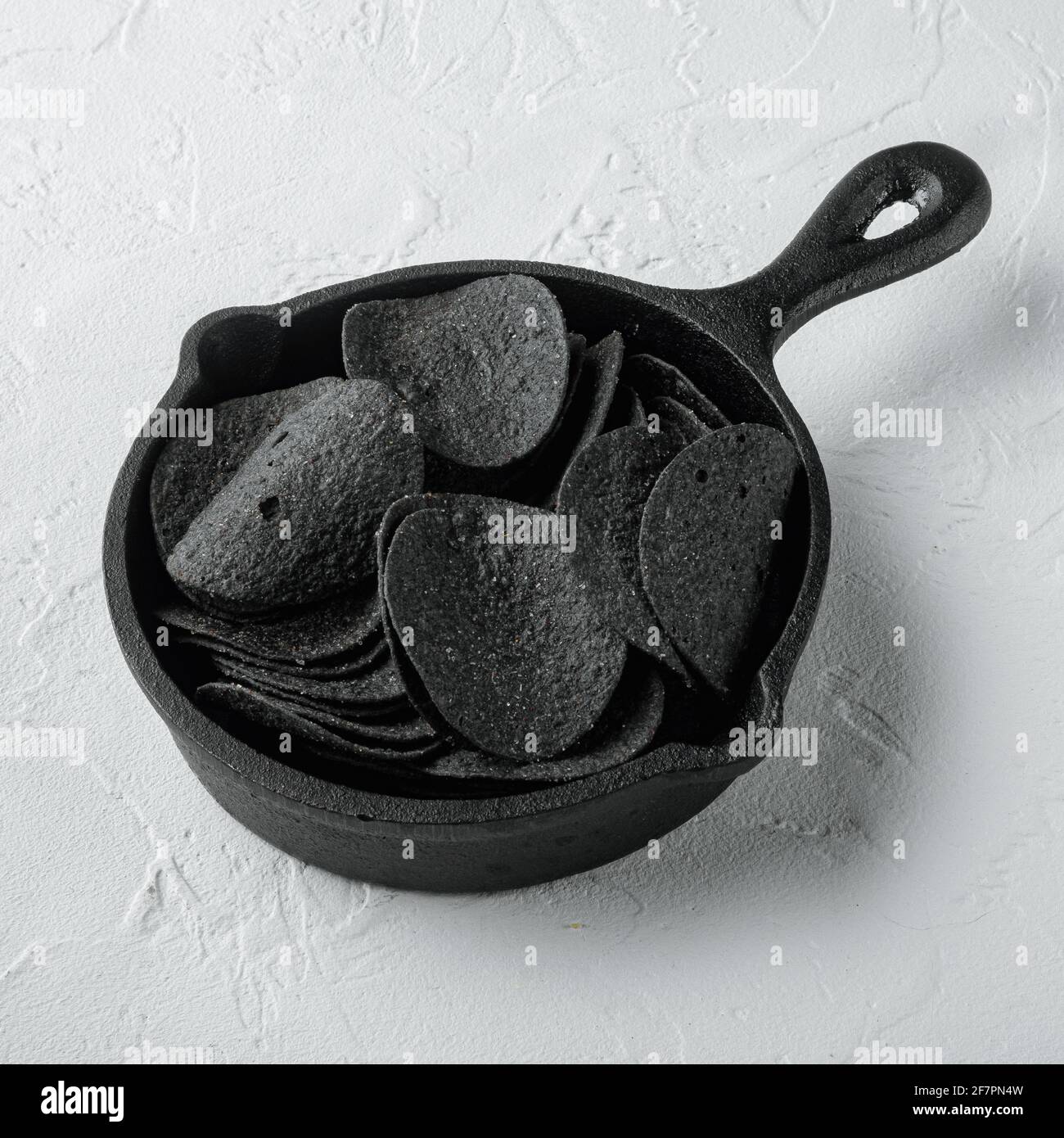 Black crispy potato chips set, in cast iron frying pan, on white stone surface, square format